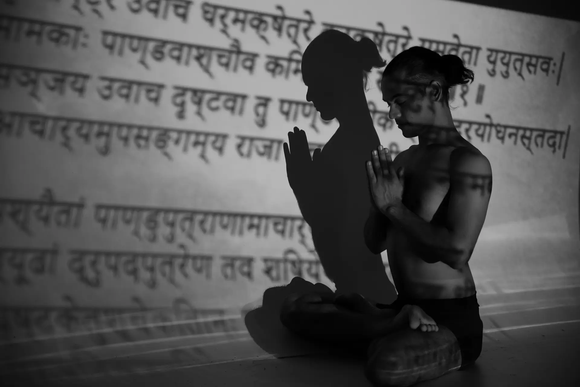 A student seated in prayer against a wall lit with projected Sanskrit verse